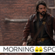Andor, clad in his leather jacket, walks in front of a red brick building.